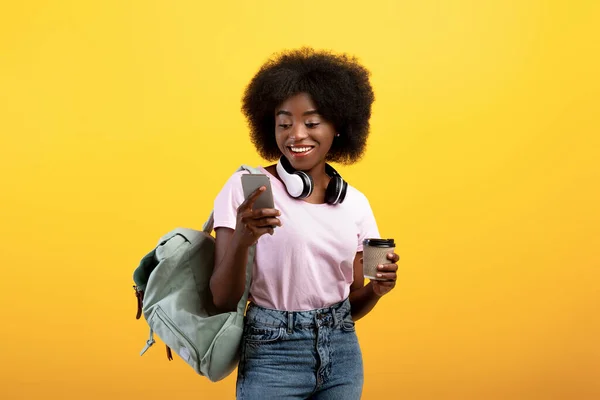 Happy black student using smartphone, wearing backpack and headphones on neck, holding coffee to go, yellow background — Stock Photo, Image