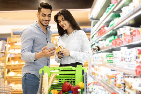 スマートフォンを使ったアラブカップル｜モダンなスーパーマーケットでの食品購入 — ストック写真