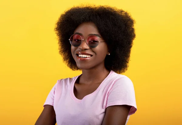 Portret van een vrolijke Afrikaans-Amerikaanse dame met een stijlvolle kleurrijke zonnebril, lachend naar de camera over een gele achtergrond — Stockfoto