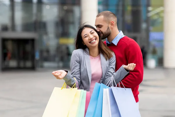 Amar chico joven besando a su novia asiática riendo con bolsas de compras, expresando afecto cerca de gran centro comercial, espacio de copia —  Fotos de Stock