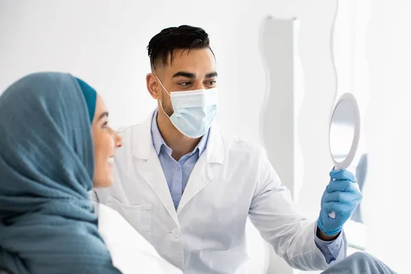 El estomatólogo árabe sostiene el espejo, mostrando el resultado del tratamiento dental a la paciente musulmana —  Fotos de Stock