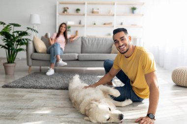 Yakışıklı Arap adam evde köpekle oynuyor, karısı arka planda kahve içiyor, boş yer var.