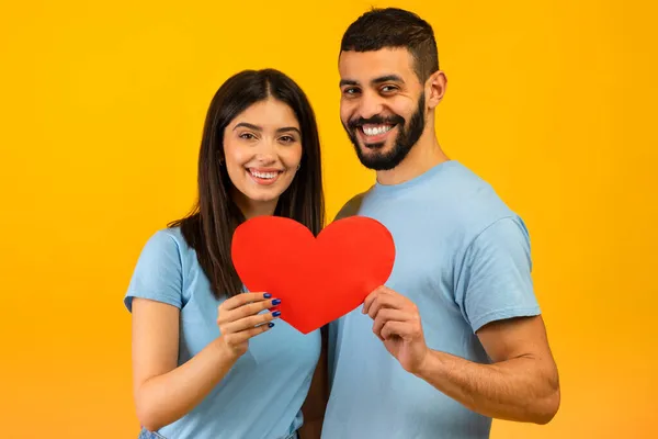 Día de San Valentín. Amantes cónyuges árabes abrazando y sosteniendo el corazón, sonriendo a la cámara sobre el fondo amarillo del estudio — Foto de Stock