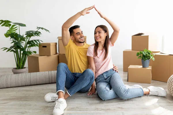 Feliz pareja internacional milenaria haciendo gesto de casa, sentado en el suelo entre cajas de cartón, mudándose a un nuevo apartamento —  Fotos de Stock