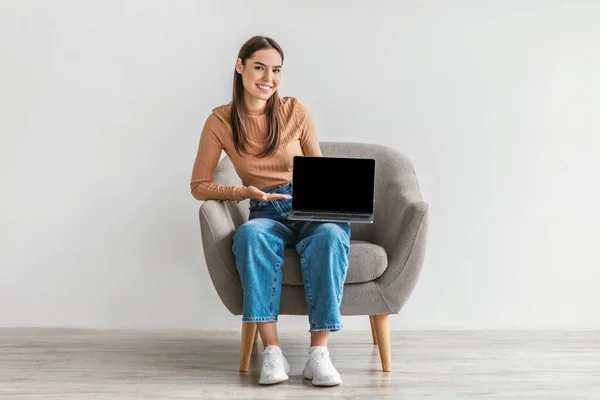 Joyeux jeune femme démontrant ordinateur portable avec écran vide, assis dans un fauteuil contre un mur blanc, maquette pour site Web — Photo