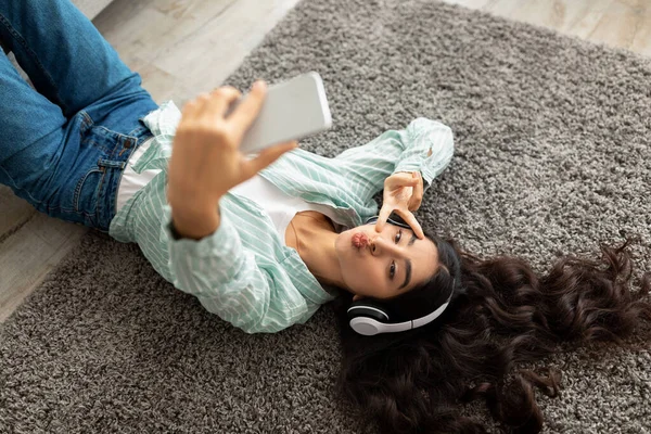 Вид сверху симпатичной индийской дамы, делающей селфи на смартфоне, лежащей на полу в наушниках, слушающей музыку дома — стоковое фото