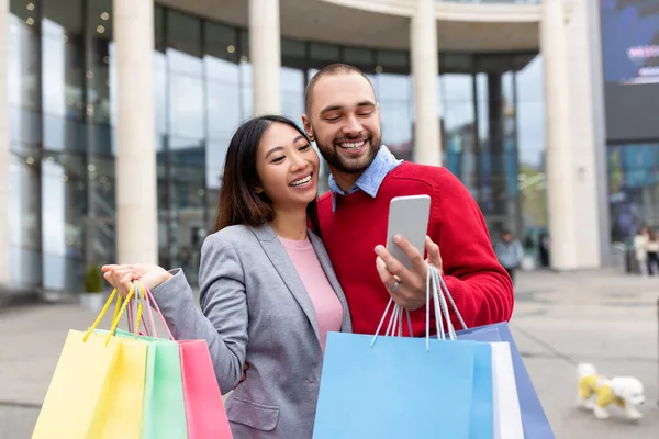 Casal multirracial positivo com sacos de presente brilhante e telefone celular colocando ordem on-line perto do shopping — Fotografia de Stock