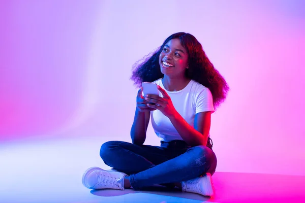 Mladá černoška sedící se zkříženýma nohama s chytrým telefonem, procházení sociálních médií, posílání zpráv přátelům v neonovém světle — Stock fotografie