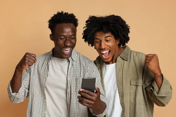 Ganar en línea. Dos chicos negros emocionados celebrando el éxito con el teléfono inteligente —  Fotos de Stock