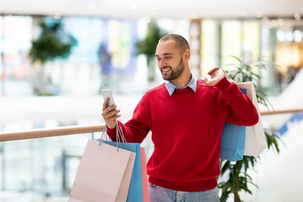 Positieve jonge man met geschenkzakjes met behulp van mobiele telefoon in het winkelcentrum, het nemen van selfie of foto, met video bellen, vrije ruimte — Stockfoto