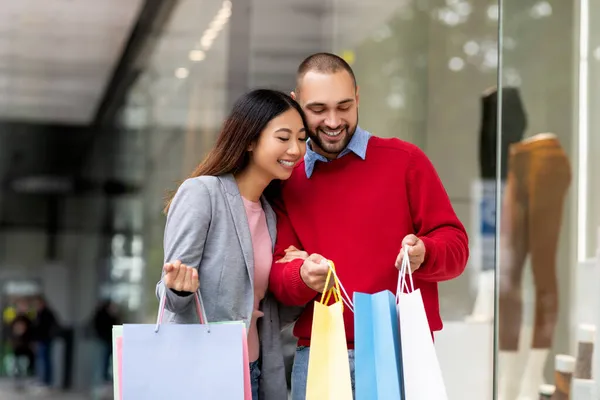 Gelukkig jong multiraciaal paar op zoek in shopper tassen, tevreden met hun aankopen na het winkelen in het winkelcentrum — Stockfoto