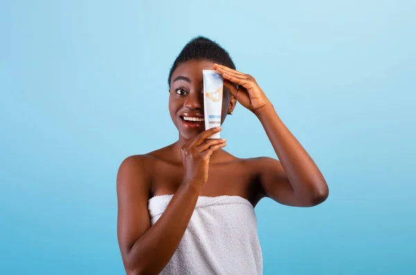Glad afrikansk amerikansk dam håller röret med kosmetisk kräm nära ansiktet, ler mot kameran över blå bakgrund — Stockfoto