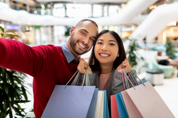 Belo casal interracial alegre segurando sacos de papel, tirando selfie no shopping, indo às compras juntos — Fotografia de Stock