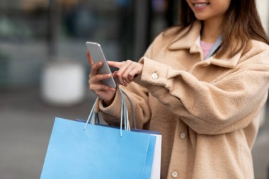 Akıllı telefon kullanan, elinde kese kâğıdı tutan, açık havada alışveriş yapan genç bir kadın.