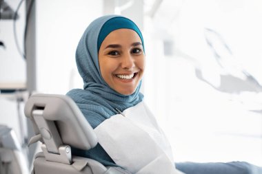 Gülümseyen Müslüman kadın hasta diş hekimi koltuğunda oturuyor, sağlık kontrolü bekliyor.