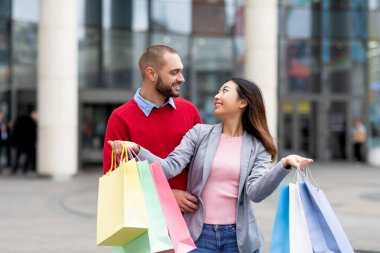 Alışveriş merkezinin yanında durup alışveriş torbalarıyla birbirlerine bakan, alışverişlerden memnun, sevgi dolu ırklar arası bir çift.