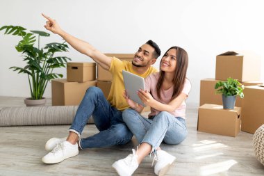 Tabletli, kutuların arasında oturmuş, yeni bir daireye mobilya yerleştirmeyi planlayan mutlu, ırklar arası bir çift.