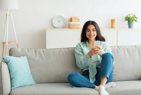 Encantadora jovem indiana sentada no sofá com smartphone, verificando as mídias sociais, conversando online em casa — Fotografia de Stock