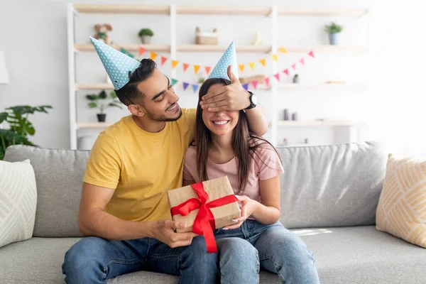 Hezký mladý Arab chlap zakrývá své přítelkyně oči, dělá narozeninové překvapení, dává jí zabalený dárek doma — Stock fotografie