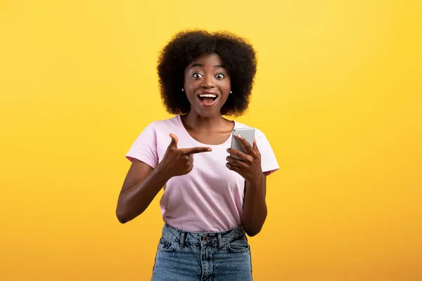 Mobiles Angebot. Fröhliche schwarze Dame zeigt auf Smartphone und blickt mit offenem Mund in die Kamera, gelber Hintergrund — Stockfoto