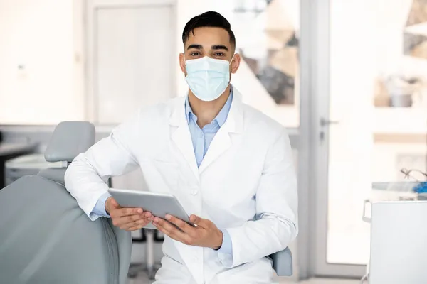 Joven médico estomatólogo árabe usando tableta digital en el lugar de trabajo en la clínica —  Fotos de Stock