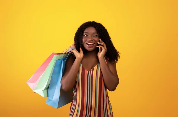 かなりアフリカ系アメリカ人の女性が買い物袋を持って携帯電話で話してオンラインショッピングの注文を確認 — ストック写真