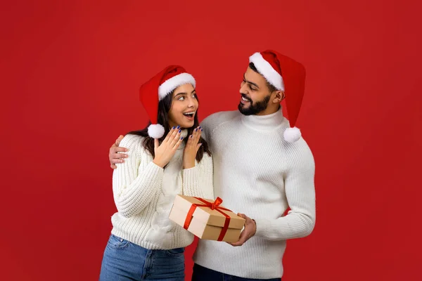 Christmas gifts. Loving arab husband surprising young wife, giving wrapped present box and embracing, red background — Stock Photo, Image