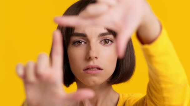 Junge Frau blickt Kamera durch Fingerrahmen, gelber Hintergrund — Stockvideo