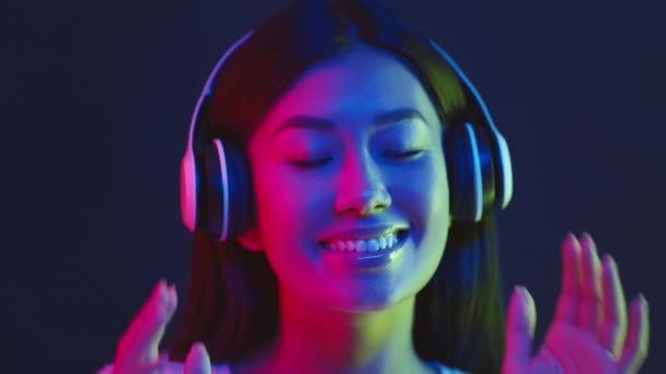 Moderne muziek. Jonge Aziatische dame draagt draadloze headset genieten van dynamische muziek, dansen in neon lichten over donkere studio — Stockvideo