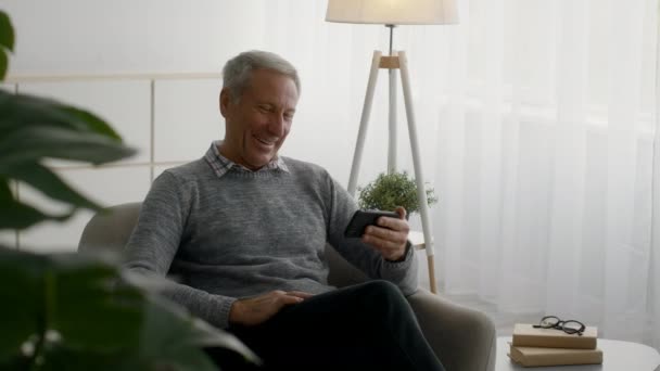 Sorrindo homem idoso relaxando em casa com smartphone, assistindo vídeos engraçados online — Vídeo de Stock