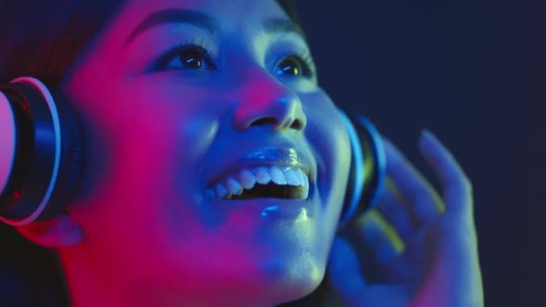 Conceptul muzicii futuriste. Tânără femeie asiatică fericită care se bucură de muzică cu căști wireless, zâmbind în lumini luminoase de neon — Videoclip de stoc