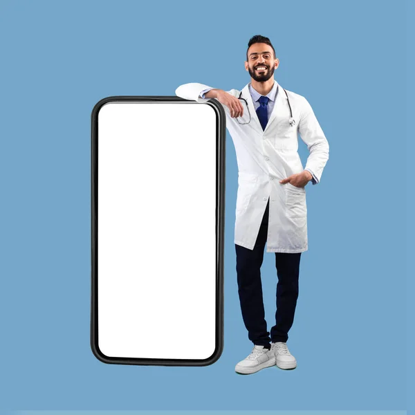 Ближневосточный доктор опираясь на большой смартфон, синий фон, площадь — стоковое фото