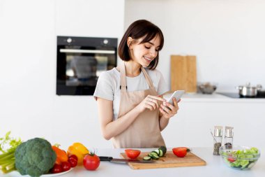 Genç bayan akıllı telefon kullanıyor ve taze salata pişiriyor. İnternette yeni tarifler arıyor. Mutfakta duruyor.