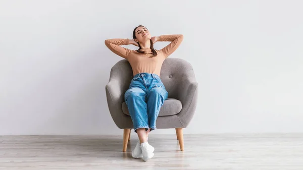 Mujer joven y pacífica sentada en un sillón, cogida de la mano detrás de la cabeza, relajante, teniendo un descanso contra la pared blanca del estudio —  Fotos de Stock