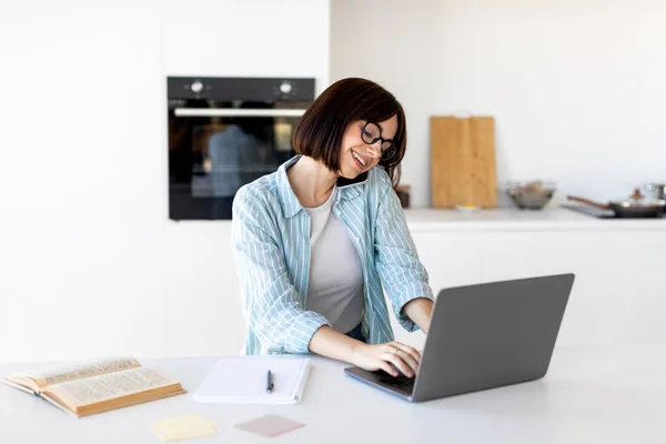 Trabalho de gerente, trabalhar remotamente. Jovem feliz falando por celular e digitando no computador portátil, sentado na cozinha — Fotografia de Stock