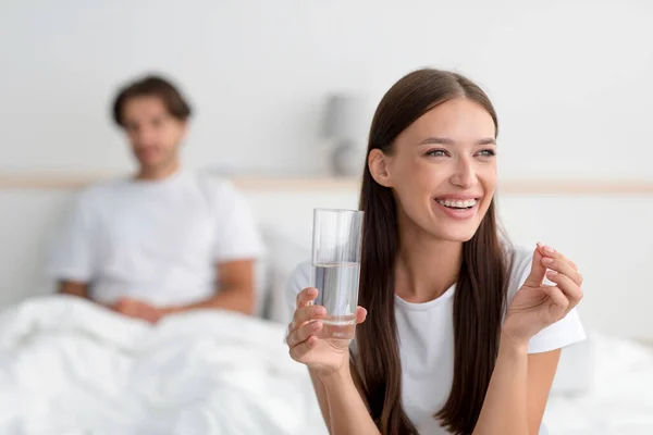 Gülümseyen Avrupalı kadın elinde bir bardak su ve hap tutarken kocası yatak odasında iç tarafta oturuyor. — Stok fotoğraf