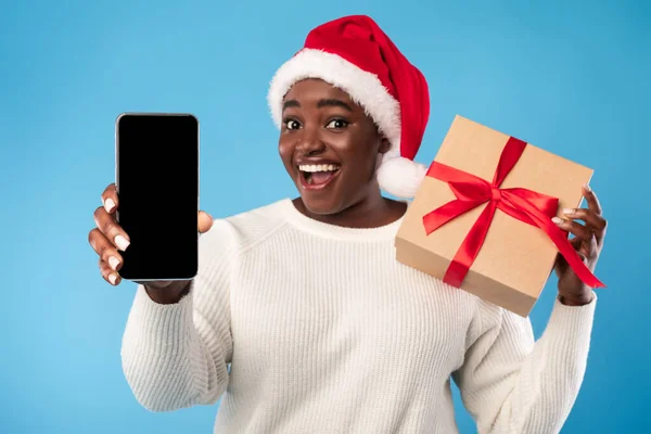 African Woman Showing Smartphone Screen Holding Christmas Gift, Blue Background — Stock Photo, Image