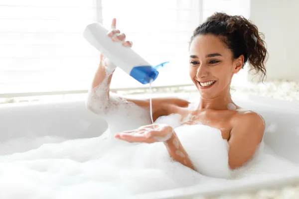 O femeie tânără fericită care face baie cu bule, turnând șampon din sticlă în mână, bucurându-se de procedura spa domestică — Fotografie, imagine de stoc