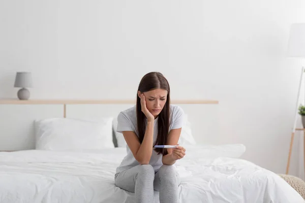Triste déception jeune femme européenne tenir test de grossesse, assis sur le lit seul dans la chambre — Photo