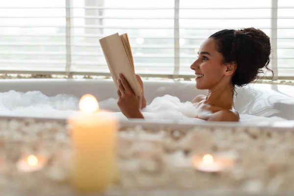 Bella giovane donna sdraiata in bagno schiumoso, libro di lettura in atmosfera rilassante con candele, all'interno. Copia spazio — Foto Stock