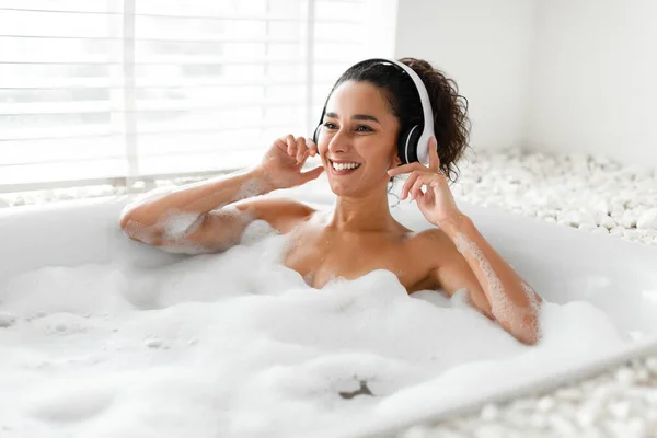 自宅で熱い泡風呂に横たわっている間に音楽を聞いてヘッドフォンで魅力的な若い女性,フリースペース — ストック写真