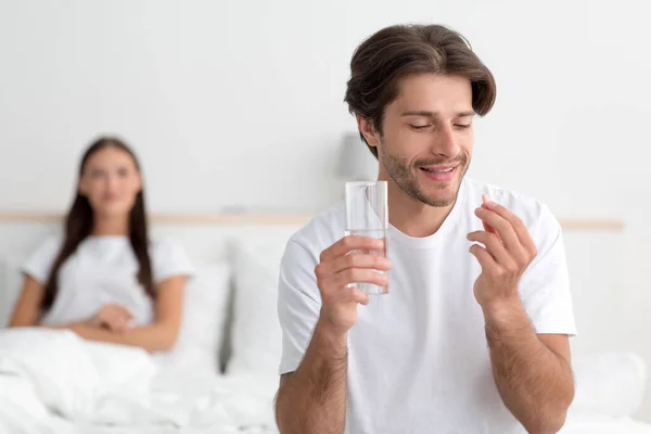 Mosolygó fiatal kaukázusi férj kezében pohár víz és tabletta, feleség vár férfi az ágyon a hálószobában belső — Stock Fotó