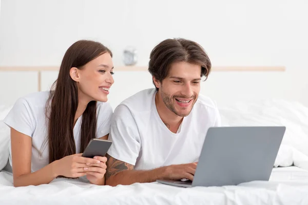 Пара юропіанців, що посміхається, лежить на ліжку, з ноутбуком і смартфоном мають онлайн дзвінок, переглядають відео — стокове фото