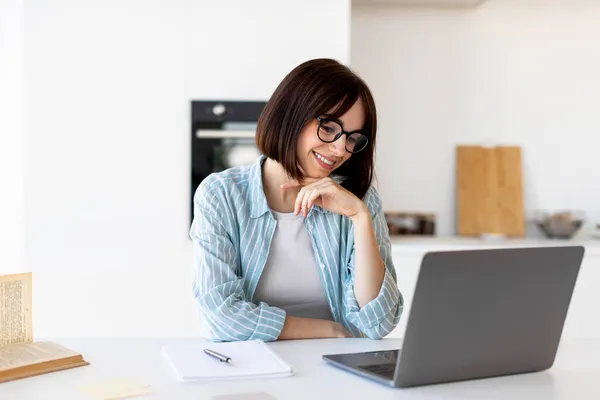 Il concetto di lavoro freelance. Felice signorina seduta a tavola con computer portatile in cucina, guardando lo schermo e sorridendo — Foto Stock