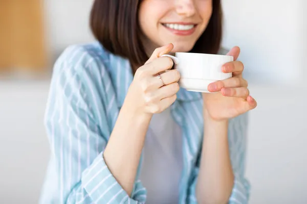Mujer joven caucásica sosteniendo taza de bebida caliente, café o té, disfrutando del aroma mientras desayuna en la cocina — Foto de Stock