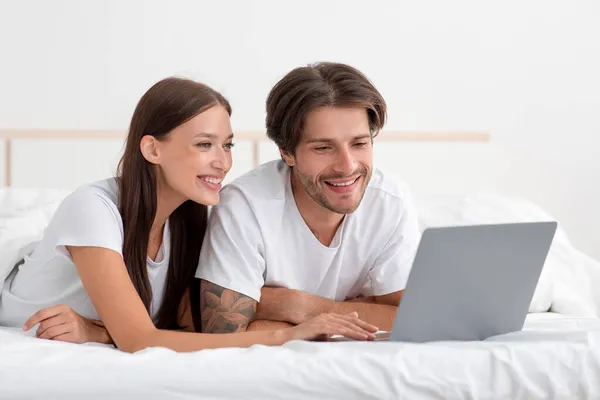 Joyeux jeune couple caucasien allongé sur le lit avec ordinateur portable ont appel vidéo, regarder la vidéo, bavarder, recherche — Photo