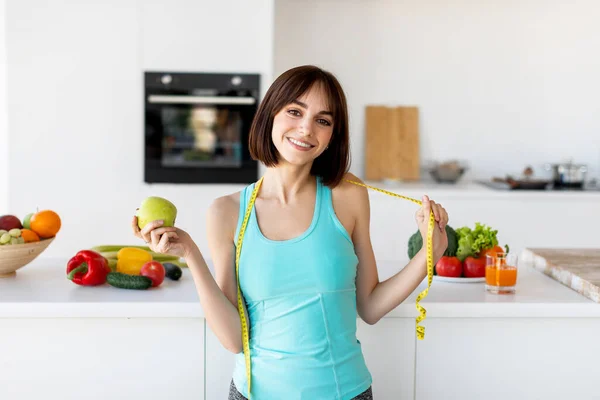 Dietní koncept. Happy fit dáma s měřicí páskou kolem krku drží zelené čerstvé jablko a usmívá se v kuchyni — Stock fotografie