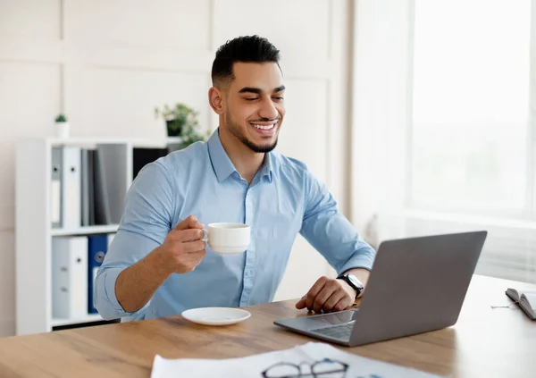 Glücklicher junger arabischer Mann trinkt Kaffee, während er im modernen Büro Laptop benutzt — Stockfoto