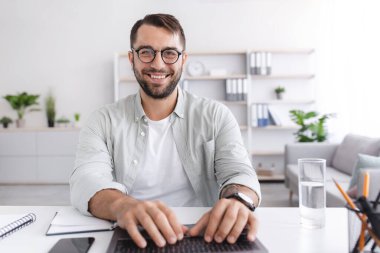 Gözlüklü neşeli, olgun, beyaz adam dizüstü bilgisayarda çalışıyor ya da oturma odasının içinde online toplantı yapıyor.