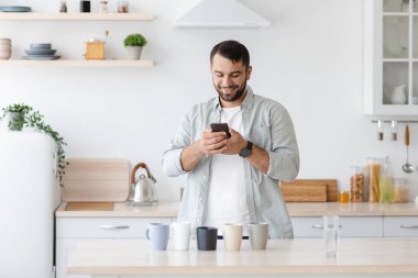 Gülümseyen orta yaşlı beyaz erkek, minimalist mutfakta çay içerken pek çok farklı fincanın fotoğrafını çeker.